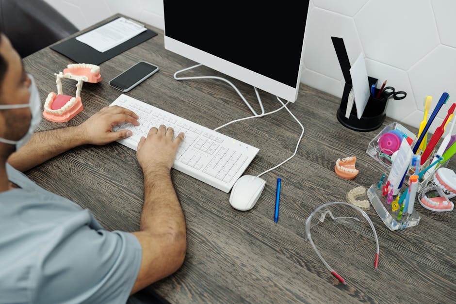 Dentist at desk typing on computer with dental models and tools around.